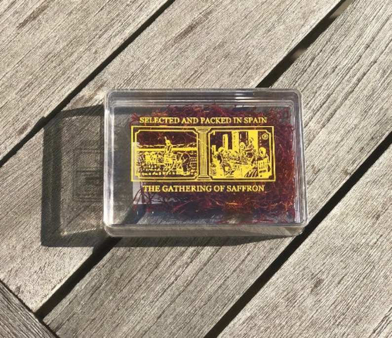 May include: A clear plastic box containing red saffron threads. The box has a gold label with the text "SELECTED AND PACKED IN SPAIN" and "THE GATHERING OF SAFFRON".