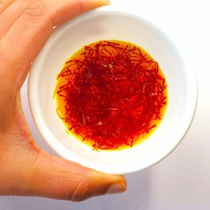 May include: A white bowl filled with saffron threads in a yellow liquid. The bowl is being held by a hand.