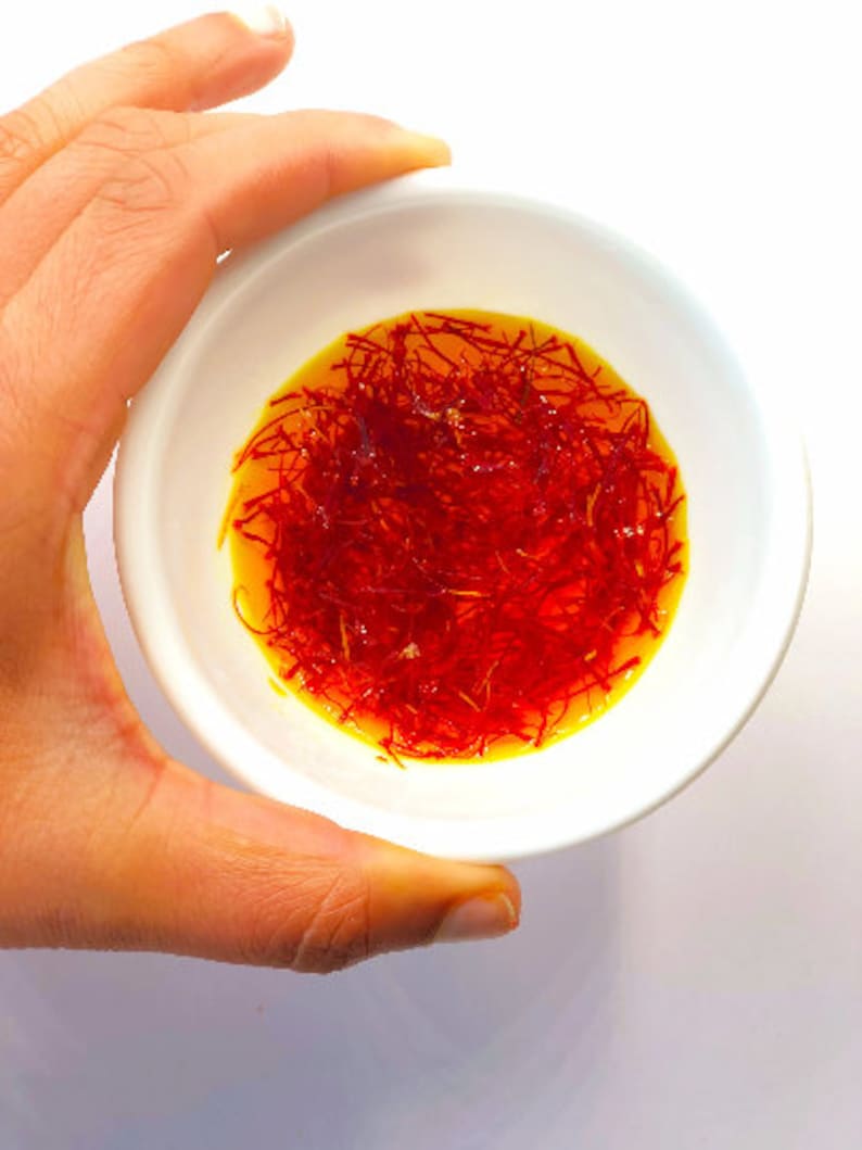 May include: A white bowl filled with saffron threads in a yellow liquid. The bowl is being held by a hand.