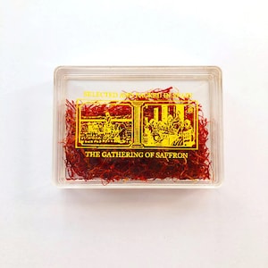 May include: A clear plastic container filled with red saffron threads. The container has a gold label that reads "Selected and Packed in Spain" and "The Gathering of Saffron".