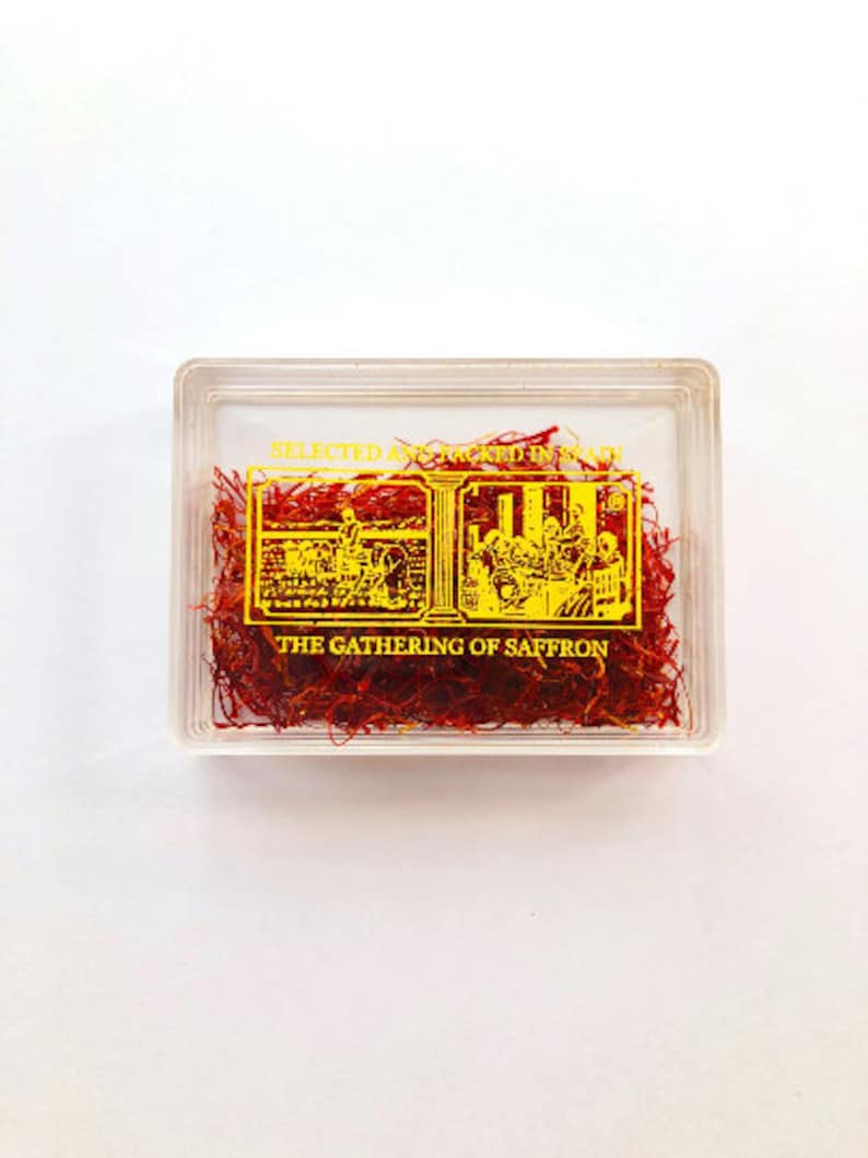 May include: A clear plastic container filled with red saffron threads. The container has a gold label that reads "Selected and Packed in Spain" and "The Gathering of Saffron".