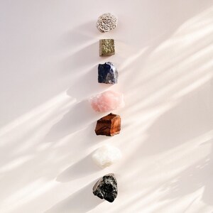 May include: A vertical arrangement of various crystals and stones on a light pink background. The crystals include a light brown selenite, a dark green pyroxene, a dark blue sodalite, a light pink rose quartz, a dark brown tiger's eye, a white selenite, and a black obsidian.