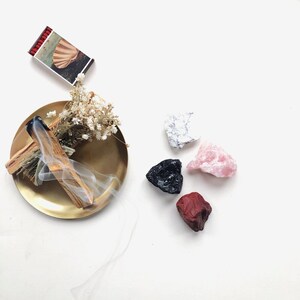 May include: A gold dish with a smoldering palo santo stick, surrounded by five crystals: white, pink, black, and red.