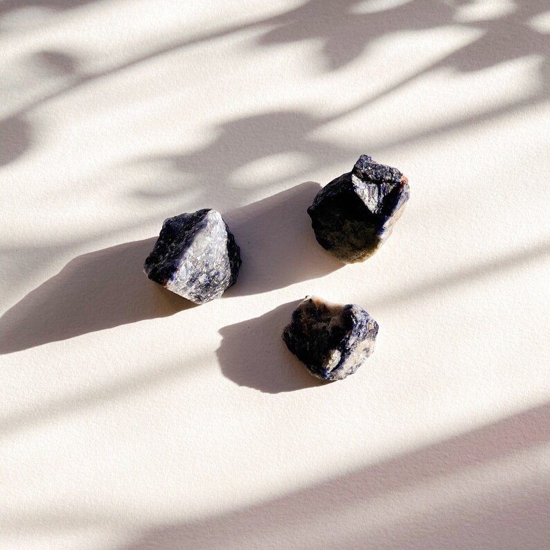 May include: Three blue and white gemstone crystals on a white surface. The crystals are rough and have a natural, unpolished look.