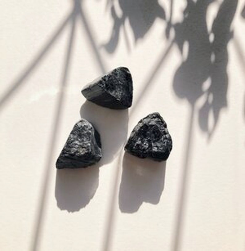 May include: Three black, rough-hewn, triangular-shaped stones. The stones are arranged in a triangular pattern on a white surface.