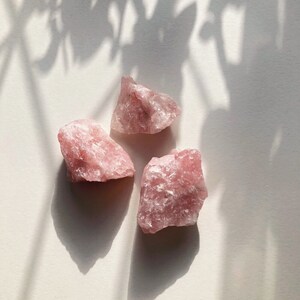 May include: Three pink rose quartz crystals on a white surface. The crystals are rough and have a natural, unpolished look.