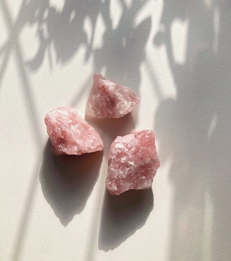 May include: Three pink rose quartz crystals on a white surface. The crystals are rough and have a natural, unpolished look.