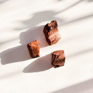 May include: Three rough-hewn, reddish-brown stones with a layered, striated texture. The stones are arranged in a triangular pattern on a white surface.