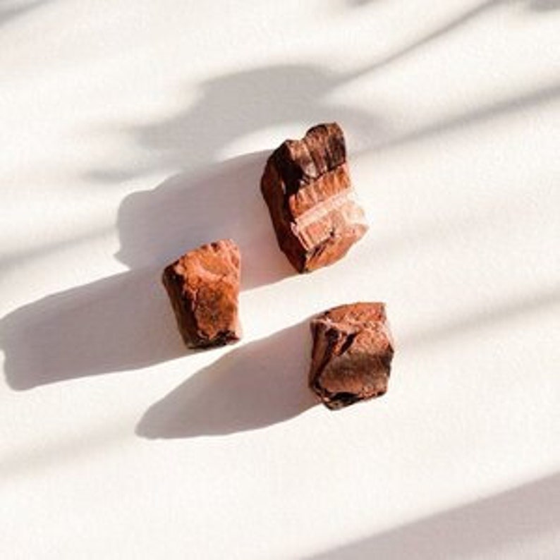 May include: Three rough-hewn, reddish-brown stones with a layered, striated texture. The stones are arranged in a triangular pattern on a white surface.