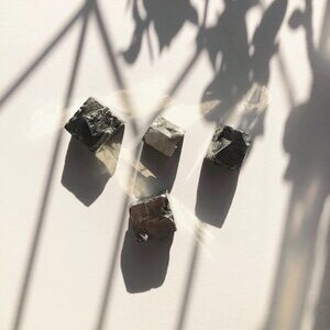 May include: Three dark gray, rough-hewn, cube-shaped crystals with a slightly iridescent sheen.