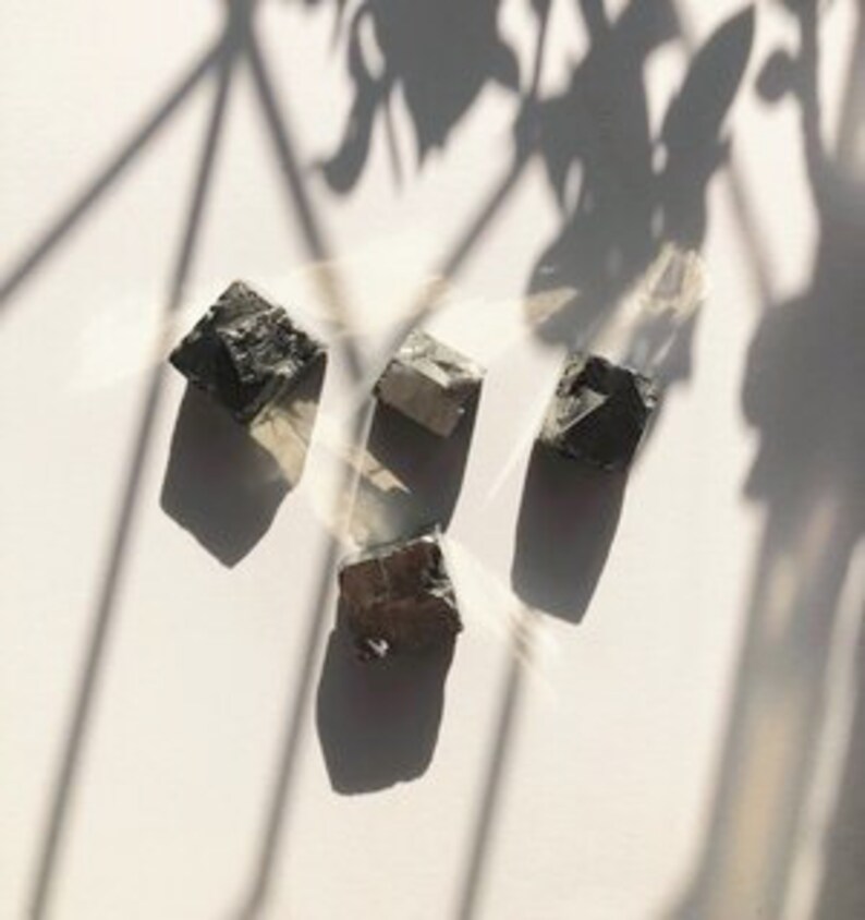 May include: Three dark gray, rough-hewn, cube-shaped crystals with a slightly iridescent sheen.