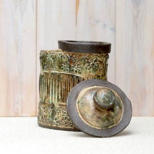 Op de afbeelding: Een keramische pot met een deksel, met een getextureerde, groene en bruine glazuur. De pot heeft een bloemenpatroon en een geribbeld patroon.