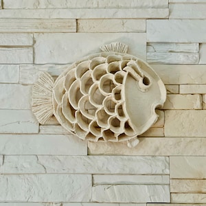 Puede incluir: Una decoración de pared de cerámica blanca en forma de pez con un diseño detallado y texturizado. El pez tiene un cuerpo redondeado y una pequeña aleta caudal.