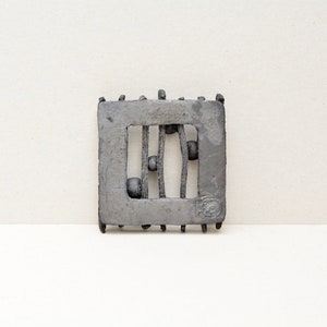 May include: A grey ceramic wall hanging with a square frame and a rectangular opening. The opening features a design of vertical lines with small round shapes.