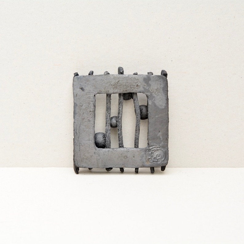 May include: A grey ceramic wall hanging with a square frame and a rectangular opening. The opening features a design of vertical lines with small round shapes.