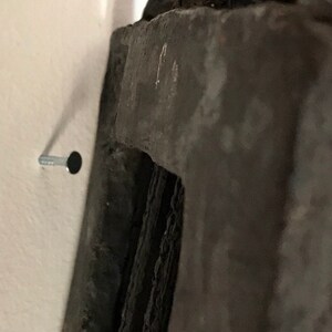 May include: A close-up of a silver nail hammered into a white wall, with a dark grey object partially visible in the background.