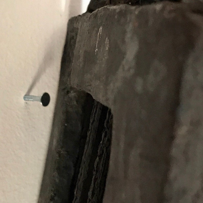 May include: A close-up of a silver nail hammered into a white wall, with a dark grey object partially visible in the background.
