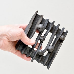 Puede incluir: Una escultura de pared de cerámica negra con forma rectangular y superficie texturizada. La escultura tiene una serie de líneas elevadas y pequeñas esferas negras redondas.