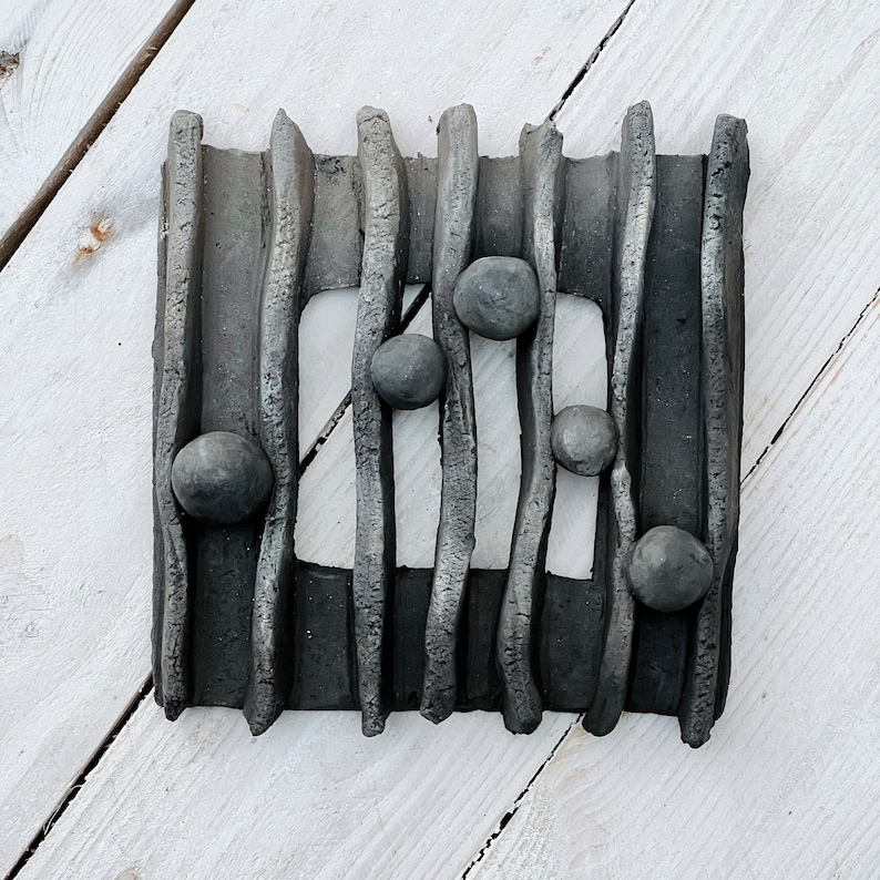 May include: Black and grey ceramic wall art with a geometric design. The art features a series of vertical lines with rounded edges and small black circles placed within the lines.