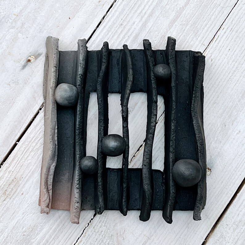 May include: Black and grey ceramic wall art with a rectangular frame and a series of black lines. The frame is decorated with small, round, black ceramic balls.