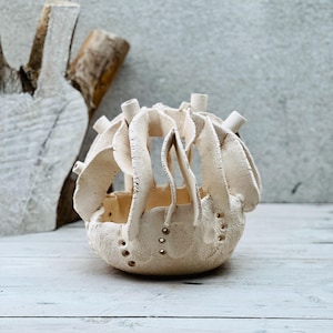 Puede incluir: Una cesta de cerámica artesanal de color blanquecino con un diseño abierto único. La cesta presenta elementos verticales y curvos y pequeñas protuberancias cilíndricas. La superficie texturizada tiene pequeños agujeros espaciados uniformemente. La cesta está sobre una superficie de madera blanca.