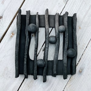 May include: Black and white abstract wall art with a rectangular frame made of thin, textured strips of clay. The frame has a cutout in the centre and is decorated with five black spheres.