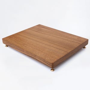 Puede incluir: Una tabla de servir rectangular de madera con un acabado natural de vetas. La tabla se eleva sobre cuatro pequeños pies redondos de color latón. La madera tiene un tono marrón cálido, sobre un fondo blanco.