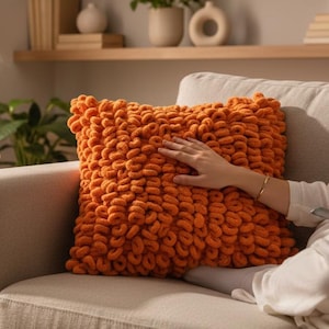 Könnte beinhalten: Ein orangefarbenes, grob gestricktes Kissen liegt auf einem beigen Sofa. Die Textur des Kissens ist deutlich sichtbar, mit einer Hand, die darauf ruht. Ein Beistelltisch mit einer weißen Tasse und ein Fenster mit transparenten Vorhängen befinden sich im Hintergrund.