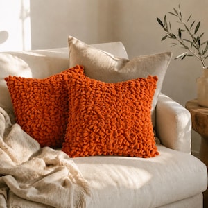 May include: Two square throw cushions featuring a vibrant orange colour and a textured, looped yarn surface. The cushions are displayed on a neutral-toned sofa alongside a plain beige cushion and a draped, light-coloured fabric throw blanket.