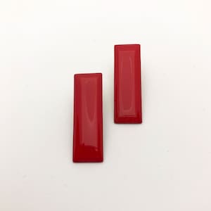 Op de afbeelding: Een paar rode rechthoekige oorbellen. De oorbellen zijn gemaakt van een glanzend, glad materiaal.
