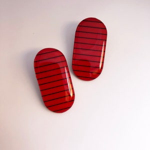 Könnte beinhalten: Zwei rote, ovale Ohrringe mit schwarzen horizontalen Streifen.