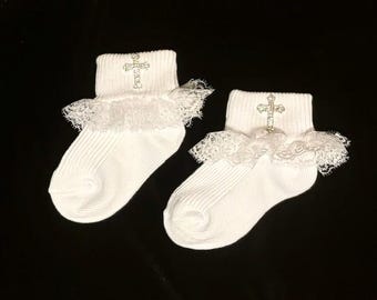 May include: A pair of white baby socks with a decorative cross and lace trim. The socks have a ribbed texture and a delicate lace ruffle around the ankle. The cross is embellished with small, sparkling details.