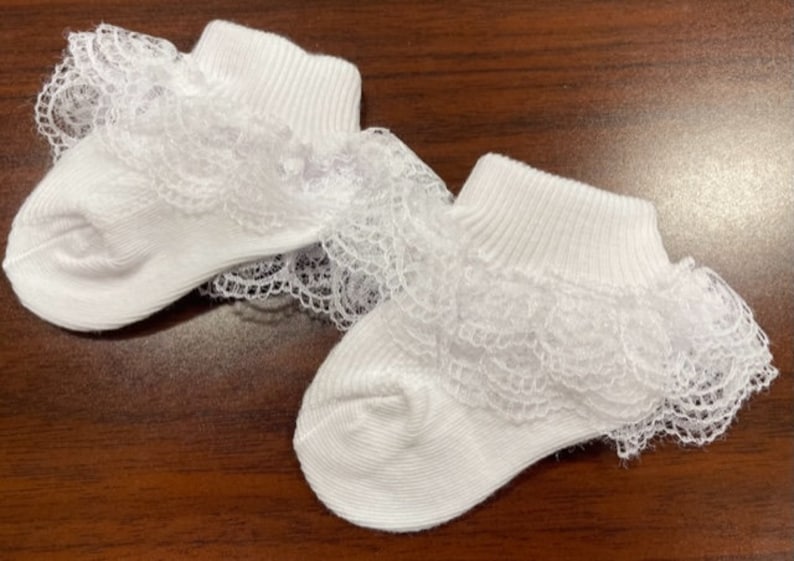 May include: A pair of white baby socks with a lace ruffle trim. The socks are made of soft cotton and are perfect for a special occasion.