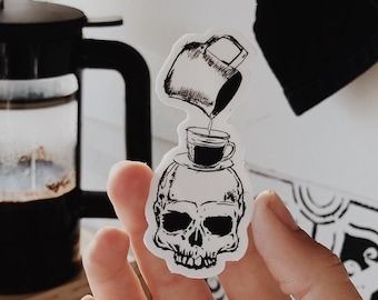 May include: A black and white sticker of a skull with a coffee cup pouring coffee into it. The sticker is on a person's hand.