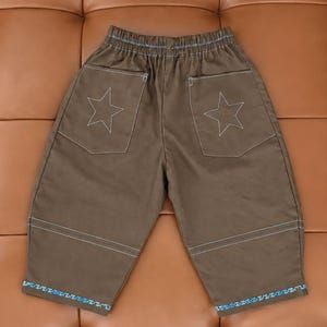 Puede incluir: Pantalones marrones con cintura elástica, dos bolsillos traseros con contornos de estrellas cosidos en blanco y detalles de costuras horizontales en azul claro sobre el dobladillo. Los bordes inferiores tienen un ribete decorativo azul y blanco.