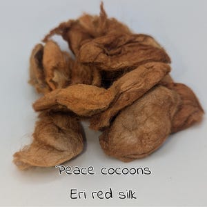 May include: A pile of Eri red silk cocoons, in shades of brown and tan, with the text "Peace cocoons" and "Eri red silk" in a handwritten font. The cocoons have a textured, fibrous appearance.