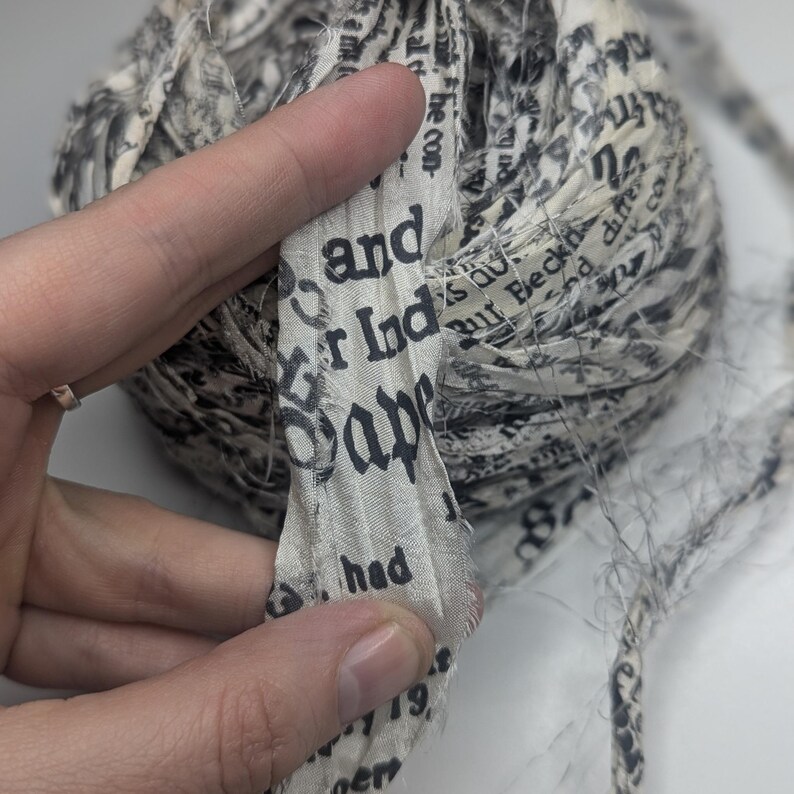Peut inclure: Une pelote de ruban de tissu imprim&eacute; de texte. Le ruban est de couleur cr&egrave;me clair avec du texte noir. Le texte comprend les mots "and", "Ind" et "Sap". Le ruban est tenu par une main.