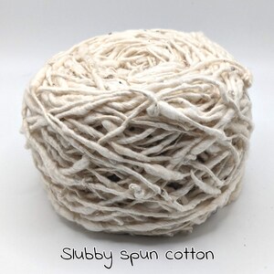 Peut inclure: Une pelote de fil de coton fil&eacute; irr&eacute;gulier, de couleur blanc cass&eacute;. Le fil a un aspect &eacute;pais et textur&eacute; avec de petites taches de fibres plus fonc&eacute;es. Le texte "Slubby spun cotton" est &eacute;crit en cursive au bas de l'image.