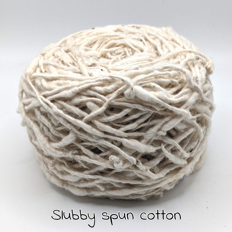 Peut inclure: Une pelote de fil de coton fil&eacute; irr&eacute;gulier, de couleur blanc cass&eacute;. Le fil a un aspect &eacute;pais et textur&eacute; avec de petites taches de fibres plus fonc&eacute;es. Le texte "Slubby spun cotton" est &eacute;crit en cursive au bas de l'image.