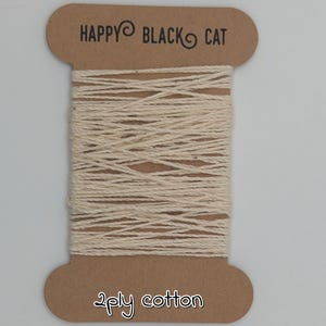 Peut inclure: Une bobine de fil de coton cr&egrave;me &agrave; 2 brins. La bobine en carton brun porte l'inscription "HAPPY BLACK CAT" et "2ply cotton". Le fil est soigneusement enroul&eacute; autour de la bobine.