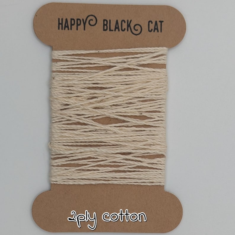 Peut inclure: Une bobine de fil de coton cr&egrave;me &agrave; 2 brins. La bobine en carton brun porte l'inscription "HAPPY BLACK CAT" et "2ply cotton". Le fil est soigneusement enroul&eacute; autour de la bobine.