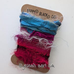 Könnte beinhalten: Eine Spule aus farbenfroher Eyelash-Sari-Seide. Die Seide ist eine Mischung aus Blau, Fuchsia und Weiß, mit einem braunen Etikett mit der Aufschrift "Happy Black Cat" und "Eyelash sari silk". Die Seide hat ein strukturiertes, gefranstes Aussehen.