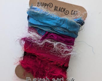 May include: A spool of colorful eyelash sari silk. The silk is a mix of blue, fuchsia, and white, with a brown tag that reads "Happy Black Cat" and "Eyelash sari silk". The silk has a textured, fringed appearance.