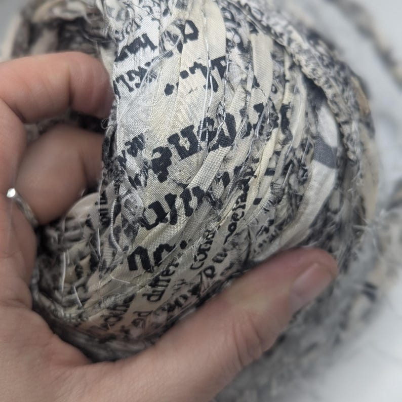 Peut inclure: Gros plan sur une boule de tissu imprim&eacute; journal. Le tissu est de couleur cr&egrave;me avec du texte noir et est &eacute;troitement enroul&eacute;. La texture semble douce et l&eacute;g&egrave;rement effiloch&eacute;e, sugg&eacute;rant un projet artisanal ou textile.