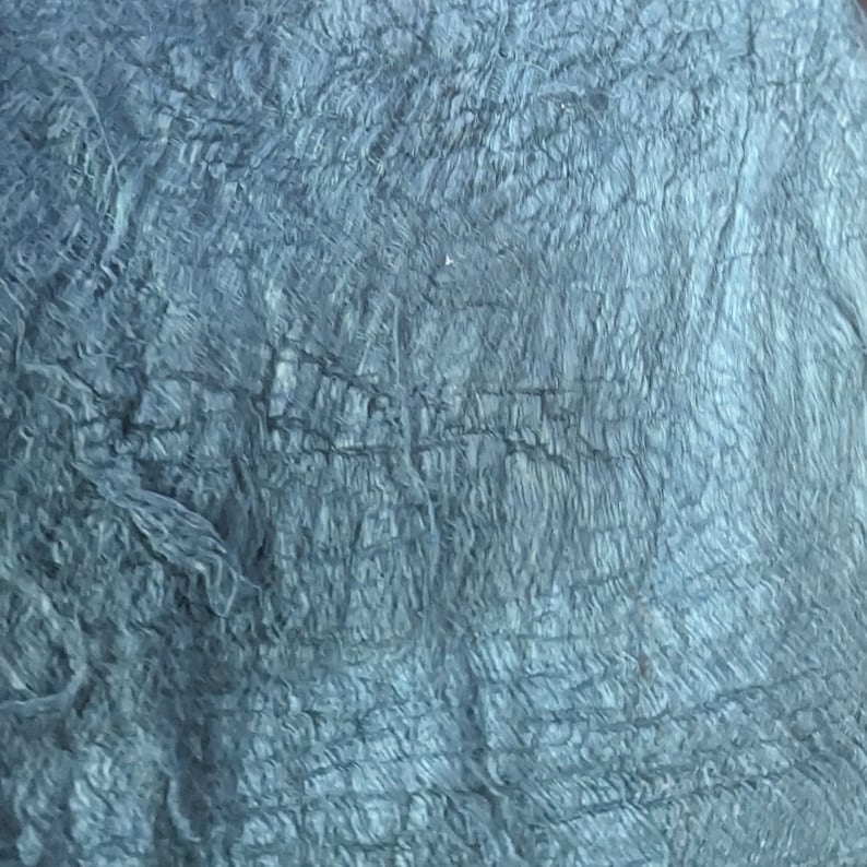 May include: A close-up of a textured, blue-grey fabric. The fabric has a layered appearance with visible ridges and a slightly rough texture. The color is a muted, cool tone, suggesting a natural material.