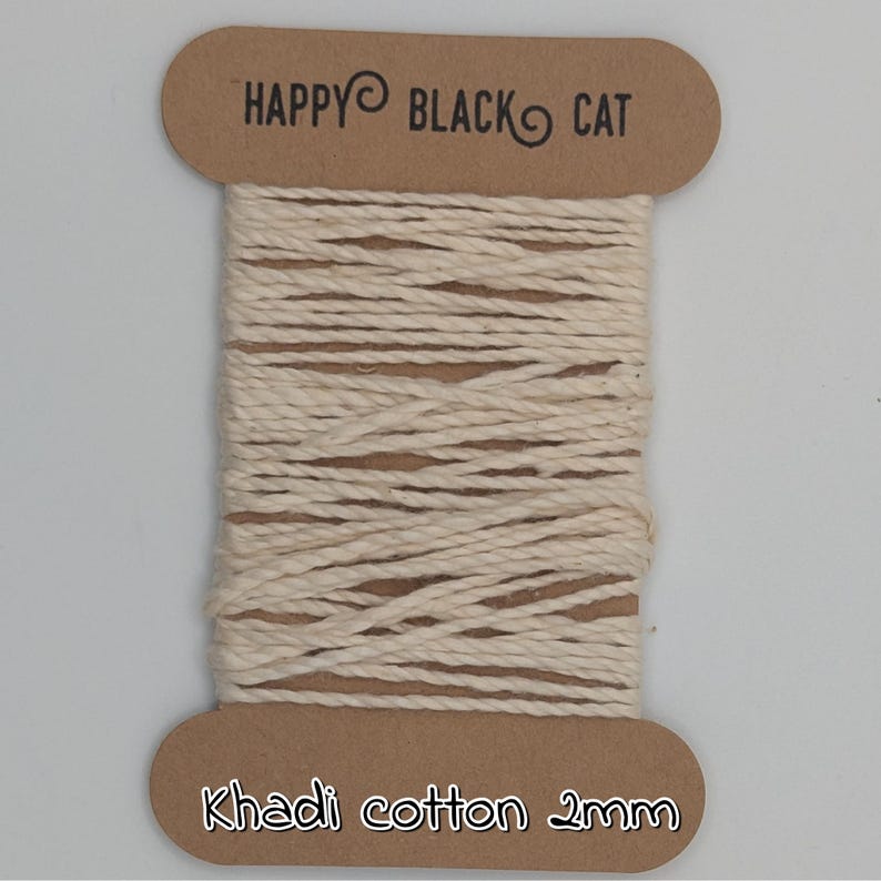 Peut inclure: Une bobine de fil de coton Khadi, 2 mm d'&eacute;paisseur, de couleur blanc cass&eacute; naturel. La bobine porte une &eacute;tiquette marron en haut avec le texte "HAPPY BLACK CAT" et une autre en bas avec "Khadi cotton 2mm".