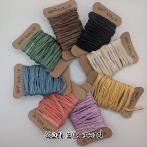 May include: A selection of sari silk cord spools in shades of green, brown, black, cream, yellow, pink, purple, and blue. Each spool is labelled with "HAPPY BLACK CAT" and the text "Sari silk cord". The spools are presented on a white surface.