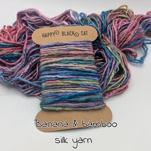 May include: A vibrant spool of silk yarn, labelled "Happy Black Cat" and "Banana & bamboo." The yarn features a blend of blues, pinks, purples, and greens. The silk yarn is wound around a cardboard spool.