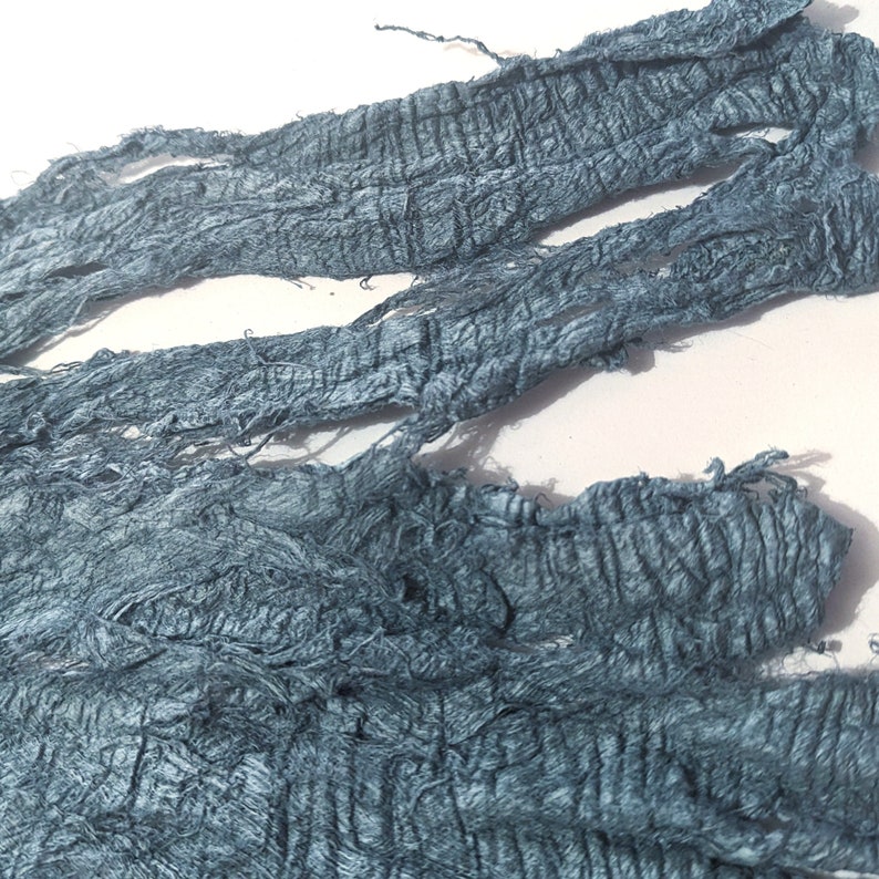 May include: Close-up of several strips of textured, teal-colored fabric. The fabric appears to be hand-dyed or distressed, with a crinkled, uneven surface. The edges are frayed, adding to the rustic aesthetic. The fabric is arranged against a white background.