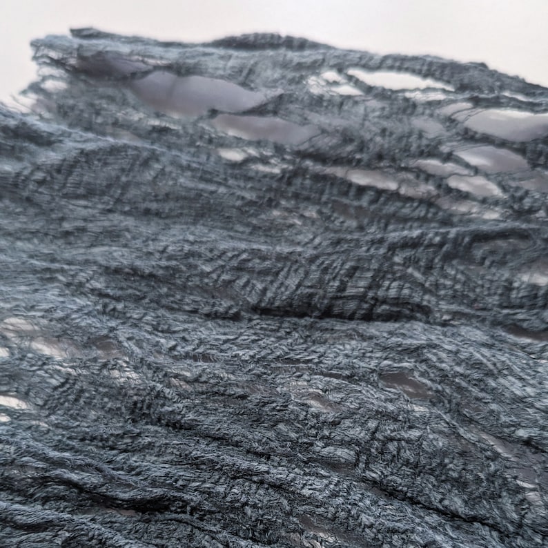 May include: Close-up of a textured, dark teal fabric with a crinkled appearance. The fabric has a distressed look with several open, elongated holes. The material appears to be lightweight and flowing, possibly used for decorative purposes.